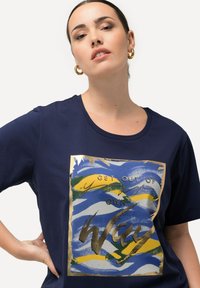 Camiseta marina de manga corta y escote redondo con un estampado abstracto colorido y texto y acentos dorados. Textura suave y lisa.
