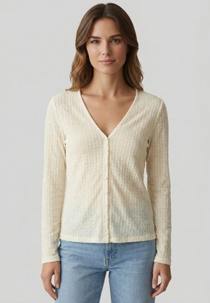 Femme portant un chemisier crème texturé à manches longues boutonné avec un col en V et un jean bleu clair, debout devant un fond uni.