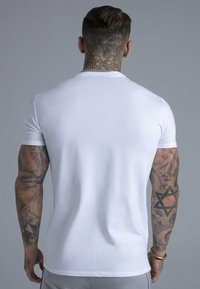 Hvid T-shirt med korte ærmer, tætsiddende design, glat bomuldstekstur, set bagfra med kroppens tatoveringer og en sølvkæde rundt om halsen.