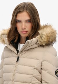 Chaqueta acolchada beige con capucha forrada de piel, diseño acolchado horizontal, cierre frontal con cremallera y detalle de logo en la manga.
