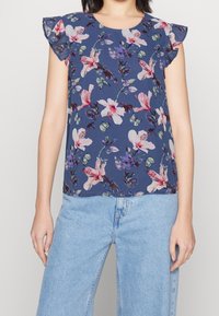 Blaues, blütenverziertes Oberteil mit kurzen, rüschierten Ärmeln, das mit rosa und weißen Blumenmustern sowie Schmetterlingen geschmückt ist. Kombiniert mit hellblauen, weiten Jeans.