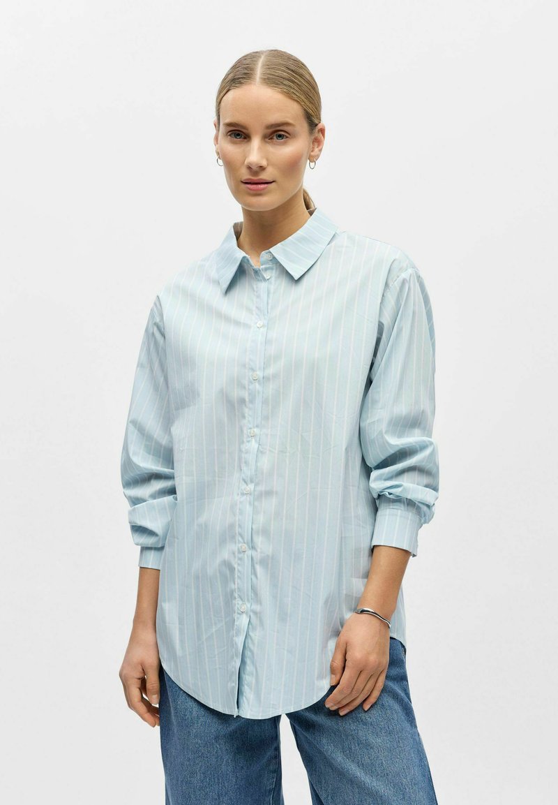 Chemise bleu clair à manches longues, boutonnée, avec des rayures blanches verticales, un col, et des manches retroussées. Dotée d’un tissu doux et d’une coupe décontractée.