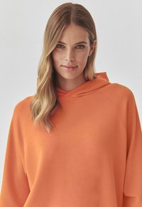 Felpa arancione con una vestibilità comoda e cappuccio. Realizzata in tessuto morbido, presenta un design semplice e una texture liscia. Nessun motivo o dettaglio accentuato.