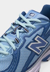Gros plan sur une chaussure de course New Balance bleu clair et bleu marine avec des lacets à motifs et une tige en tissu mesh.