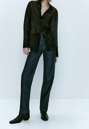 Personne portant un chemisier noir transparent à boutons avec motifs floraux, un jean droit foncé et des bottines noires sur un fond uni.