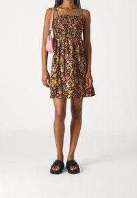 Frau trägt ein ärmelloses Blumenkleid mit einem gerafften Oberteil, schwarze Sandalen und eine kleine rosa Umhängetasche.