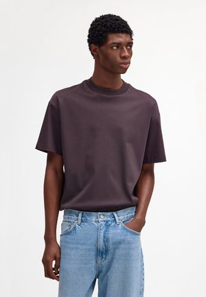 Giovane uomo con capelli corti e ricci che indossa una t-shirt oversize viola scuro e jeans azzurri, che guarda di lato con una mano in tasca.