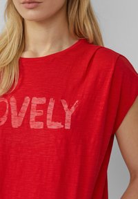 T-shirt rouge en tissu léger, avec une coupe ample et des manches courtes. L'avant affiche le mot "LOVELY" en impression blanche décolorée.