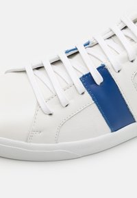 Białe skórzane sneakerzy z niebieskim akcentem, zaokrąglonym noskiem i białymi sznurowadłami. Teksturowana powierzchnia i gumowa podeszwa zwiększają przyczepność.