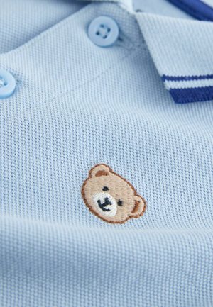 Tricou polo albastru deschis cu țesătură texturată, având două nasturi albastru deschis și un logo brodat cu urs maro pe piept.