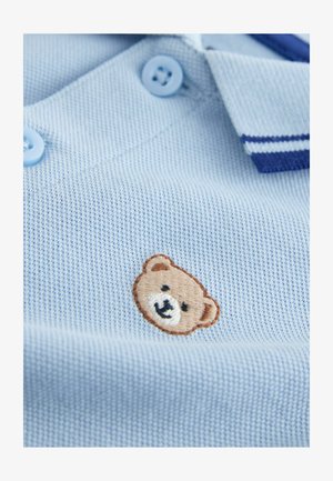 Svetlomodrá polokošeľa s textúrovanou tkaninou, ktorá má dva svetlomodré gombíky a vyšívané hnedé logo medveďa na hrudi.