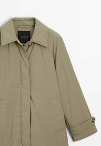 Grüne, leichte Jacke mit kragenlosem Hals, langen Ärmeln und Seitentaschen. Glatte Textur, minimalistisches Design und Knopfdetails an den Manschetten.