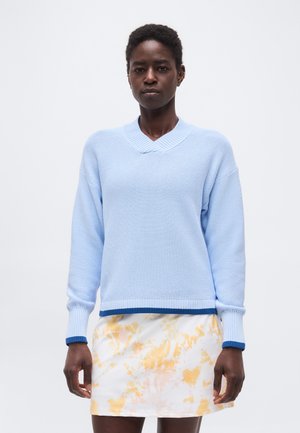 Femeie purtând un pulover bleu cu decolteu în V și margine albastru închis, și o fustă albă cu modele florale galbene, stând în fața unui fundal simplu.