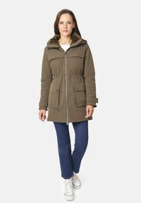 Parka de color verde oliva con cierre frontal, capucha forrada de piel y dos bolsillos frontales; llevada sobre unos vaqueros azules y zapatillas blancas.