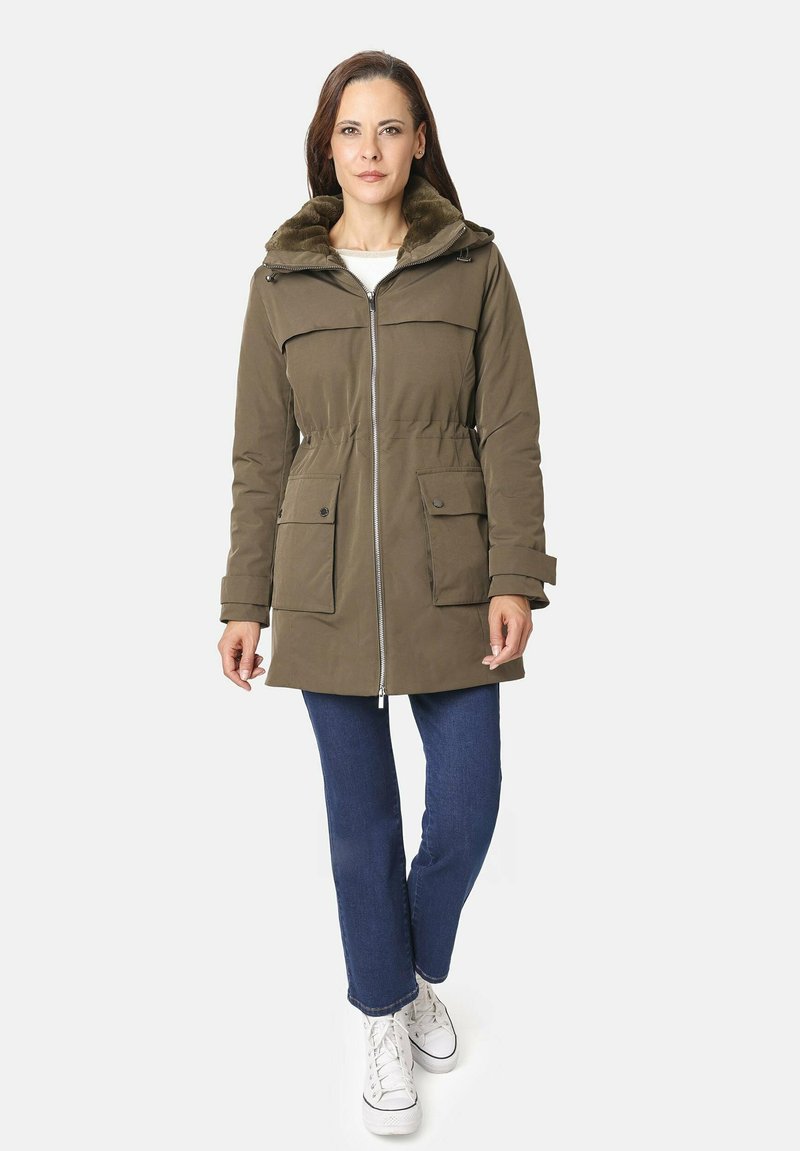 Parka de color verde oliva con cierre frontal, capucha forrada de piel y dos bolsillos frontales; llevada sobre unos vaqueros azules y zapatillas blancas.