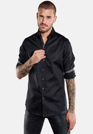 Mann mit kurzen Haaren und Tätowierungen, der ein schwarzes Langarmhemd aufknöpft, dunkle Jeans trägt und vor einem schlichten weißen Hintergrund steht.