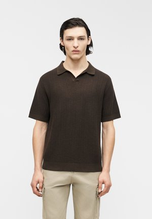 Joven con cabello oscuro, vestido con un polo de manga corta texturizado de color marrón oscuro y pantalones cargo beige claros, de pie frente a un fondo liso.