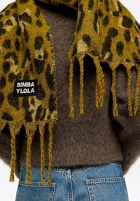 Żółto-brązowy szal w panterkę wykonany z miękkiej tkaniny, z frędzlami na brzegach i czarną metką z napisem „BIMBA YLOLA”.