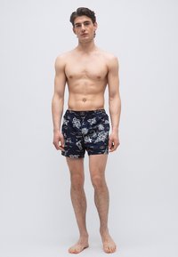 Νέος άνδρας όρθιος με γυμνά πόδια φορώντας μπλε σκούρο μαγιό με λευκό τροπικό σχέδιο, σε απλό γκρι φόντο.