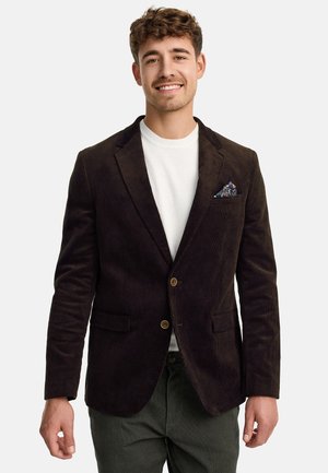 Ung mand smilende, iført en mørkebrunt kordfløjlsblazer med mønstret lommetørklæde, hvid skjorte og mørkegrønne bukser mod hvid baggrund.