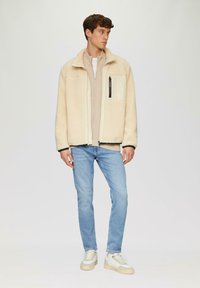 Cremefarbene Fleecejacke mit schwarzem Besatz, mit Reißverschlusstasche auf der Brust, über einem beigen Oberteil getragen, kombiniert mit hellblauen Jeans und beigen Sneakers.