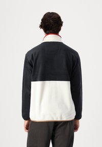 Pile pullover con sezione superiore nera, sezione inferiore bianca e colletto rosso. Presenta una texture liscia e dettagli di cuciture sottili.