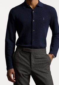 Marinblå stickad långärmad pikétröja med krage och knappstängning. Buren med grå strukturerade byxor och en klocka.