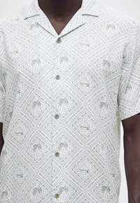 Άνδρας που φοράει πουκάμισο με κοντό μανίκι και κουμπιά, με ανοιχτό πράσινο τροπικό μοτίβο από φοίνικες και καλύβες με ψάθινη σκεπή σε λευκό ύφασμα.