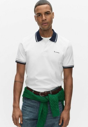 Vit pikétröja med marinblå krage och ärmdekorationer, med en liten broderad logotyp. Bärs med blå denim och en grön stickad tröja knuten i midjan.