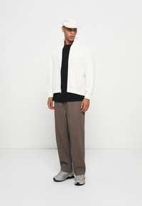 Giacca bianca con zip sopra una t-shirt nera, abbinata a pantaloni a righe marroni e sneakers grigie. Un cappellino chiaro completa l'outfit.