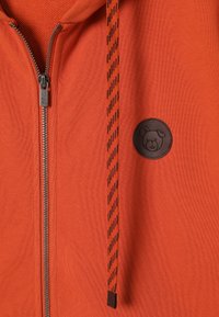 Oranje zip-up hoodie met een getextureerde stof, zwarte gestreepte trekkoorden en een ronde, bruine patch met een berenontwerp op de borst.