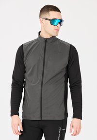 Grå ärmlös jacka med dragkedja fram, kombinerad med en svart långärmad topp. Modellen bär blåtonade pilotglasögon.