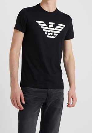 Man draagt een zwart T-shirt met korte mouwen met een wit gestileerd adelaarlogo en de letters "GA" op de borst, gecombineerd met donkergrijze spijkerbroek.