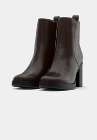 Botas de tornozelo em couro castanho escuro com fecho lateral, saltos em bloco e alças para puxar, contra um fundo claro e simples.