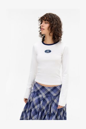 Mulher com cabelo encaracolado, vestindo uma camisa de mangas longas branca com colarinho azul-marinho e emblema, combinada com uma saia xadrez azul e cinza, de pé de lado.