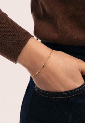 Pulsera de oro con una cadena delicada, que presenta una pequeña piedra verde y detalles de cuentas redondas; llevada en una muñeca, con un suéter marrón visible.