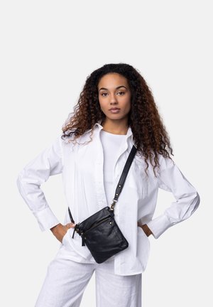 Sort læder crossbody taske med en lynlåslomme; har guld hardware og en justerbar rem, parret med en hvid skjorte og bukser.