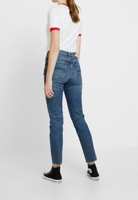 Högmidjade blå jeans med en figursydd silhuett, med fransar vid fållarna och bakfickor, kombinerat med en vit skjorta med röda detaljer.