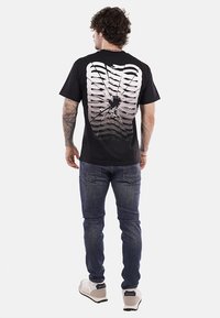 T-shirt nera con una grande grafica di una gabbia toracica bianca sul retro, caratterizzata da un design testurizzato e un effetto foro al centro.