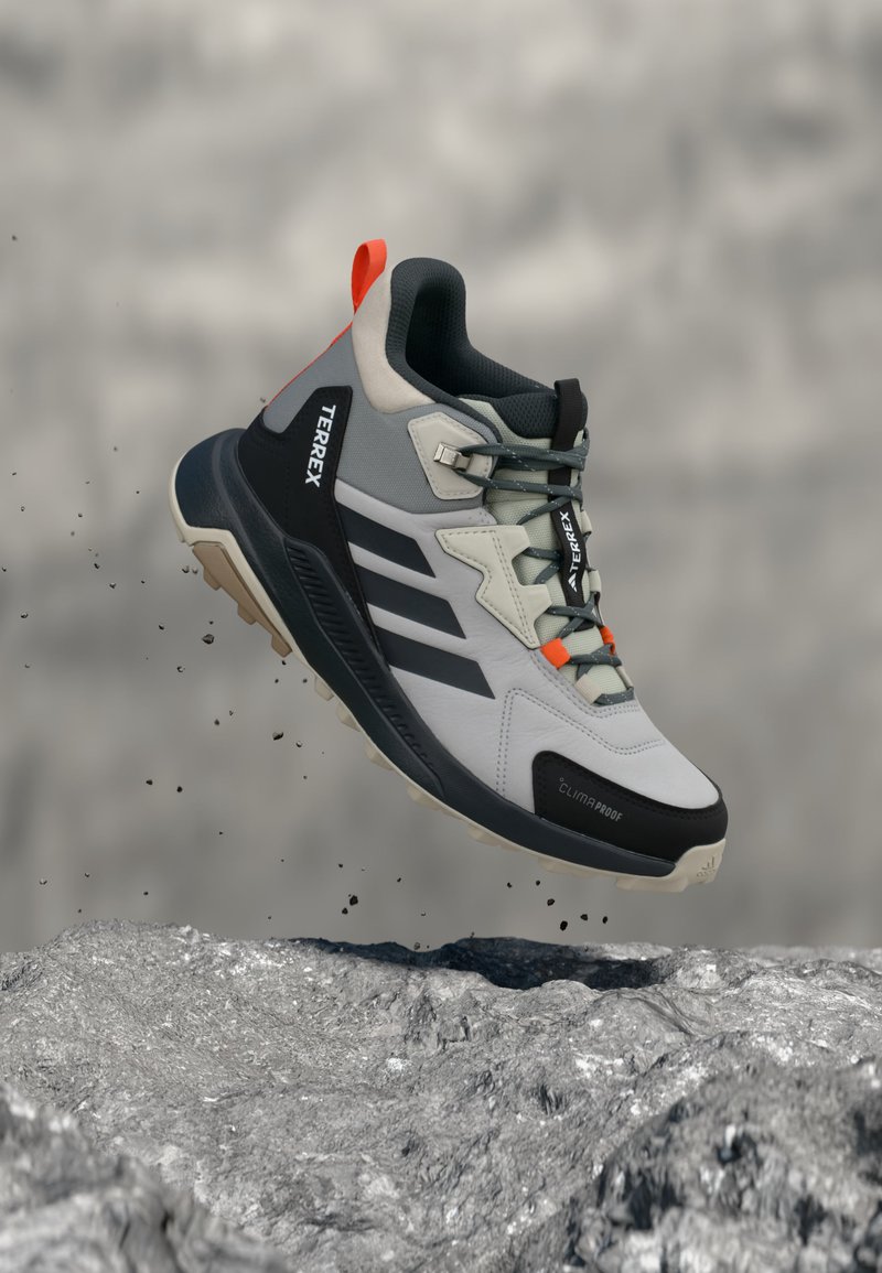 Ανοιχτό γκρι και μαύρο πεζοπορικό παπούτσι Adidas Terrex με πορτοκαλί λεπτομέρειες, κρεμασμένο πάνω από μια βραχώδη επιφάνεια, παρουσιάζοντας την ανθεκτική σόλα και την ετικέτα Climaproof.