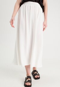 Jupe maxi blanche fluide avec une taille froncée. Tissu léger avec une texture subtile. Associée à des sandales noires ornées de clous.