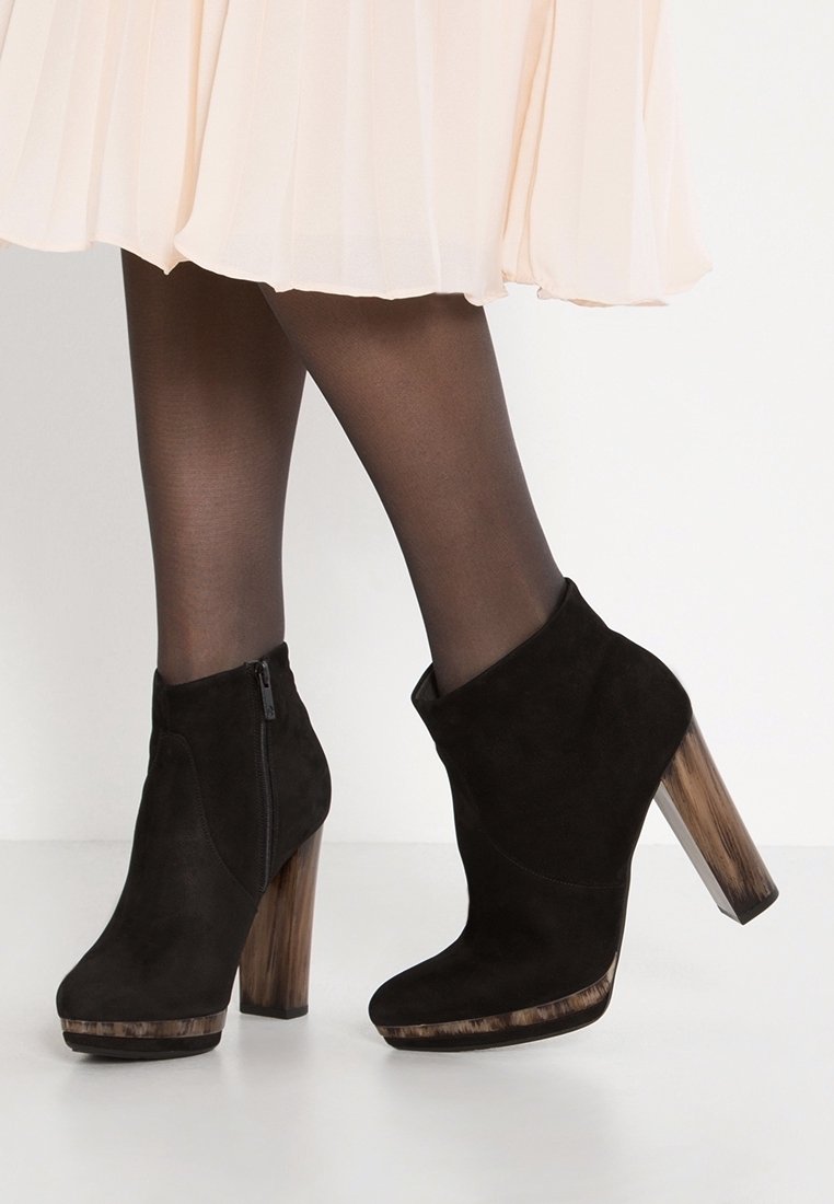 Bottines en daim noir avec talons carrés en bois, portées avec des collants noirs transparents et une jupe plissée rose pâle.