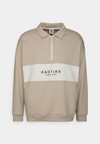Beige tröja med dragkedja i kragen, med ett vitt horisontellt rand och svart "KAOTIKO since 1999" logotyp. Mjuk, texturerad tyg.