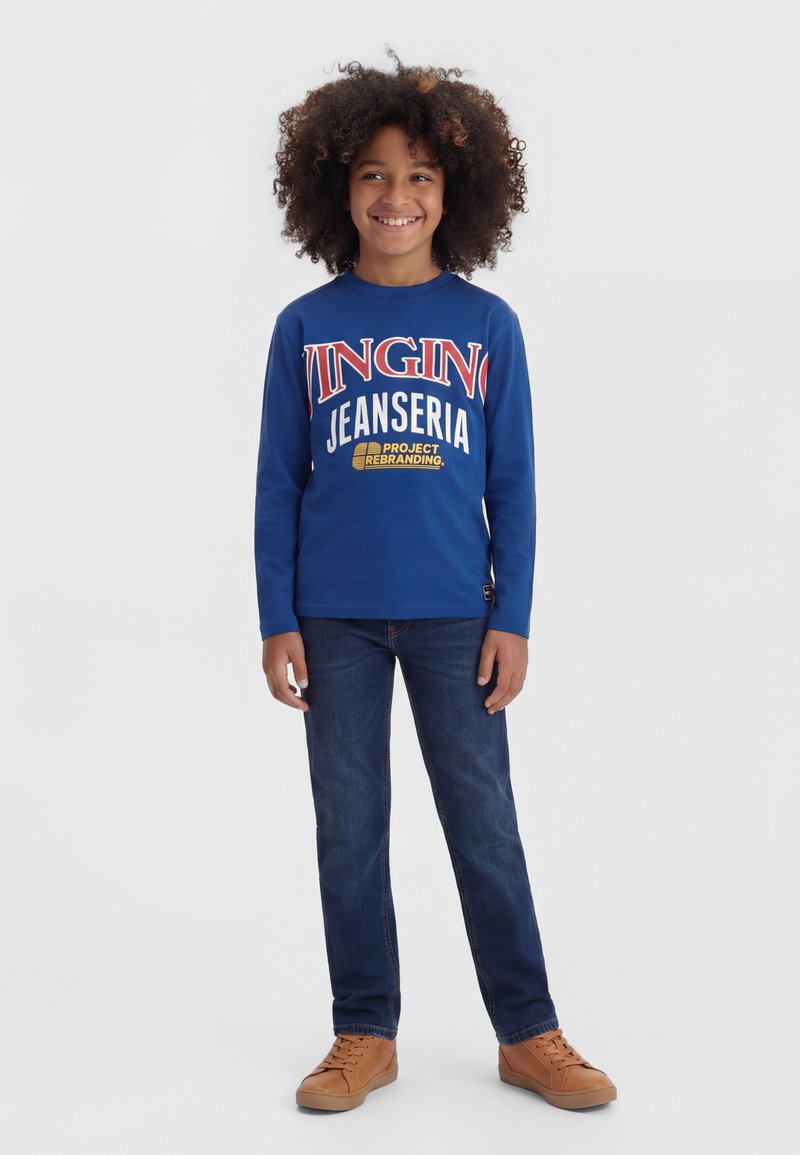 Usmievavé dieťa s kučeravými vlasmi, oblečené v modrom tričku s dlhým rukávom s textom, tmavých jeansoch a hnedých teniskách, stojace na bielom pozadí.