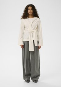 Kreemi värvi ülasärk, millel on sõlm detail vööl, koos lõtvade hallide triibuliste pükste ja sõlmedega hallide kingadega. Minimalistlik disain.