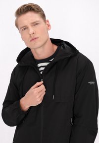 Zwarte zip-up jack met een capuchon, een voorzak en elastische manchetten. Het model draagt een marineblauwe en witte gestreepte shirt eronder.