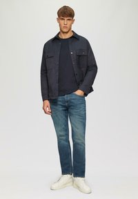 Marineblaues Hemdjackett über einem schwarzen T-Shirt, kombiniert mit hellblauen Jeans und weißen Sneakern. Glatte Stoffqualität, lässiges Design, zwei Brusttaschen.