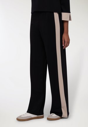 Pantaloni a gamba larga in tessuto nero, con una striscia laterale beige a contrasto. Texture liscia con cintura elastica. Indossati con sneakers casual.