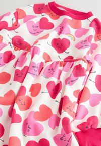 Punainen ja vaaleanpunainen sydänkuvioinen mekko valkoisella taustalla, jossa on koottu vyötärö, pitkät hihat ja ribbaus vaaleanpunaisessa kaula-aukossa.