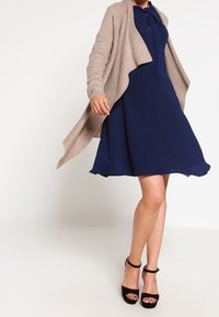 Abito blu navy con vita regolata e gonna svasata, abbinato a un cardigan aperto in maglia beige chiaro. Tacchi neri a strappi completano il look.